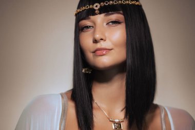 Parlak siyah saçlı, Kleopatra 'dan esinlenilmiş altın başlık takan şık bir kadın pembe bir arka plana karşı kendine güvenen bir gülümsemeyle poz veriyor. Lüks tarzı ve özgüvenli ifadesi bu görüntüyü oluşturuyor.