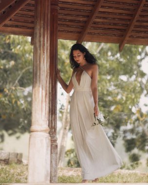 Beyaz pileli elbiseli zarif bir kadın tropikal çardağın altında çiçeklerle çıplak ayakla yürüyor. Romantik tanıtım ve editoryal kullanım için çok kaliteli bir gelin ve moda imajı..