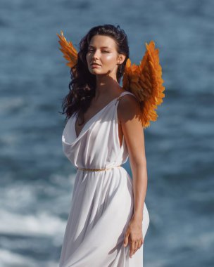Yunanca esinlenilmiş, okyanus kıyısında altın kanatlı bir elbise giyen kendine güvenen bir kadının göz kamaştırıcı yakın çekimi. Fantezi moda kampanyaları, güzellik başyazıları ve ruhani temalar için mükemmel..
