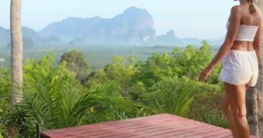 Beyaz yaz kıyafeti giymiş genç bir kadın ahşap bir platforma çıkar ve nilüfer pozisyonunda oturur, nefes kesici bir Krabi dağ zeminine karşı meditasyon yapar. Sağlık ve seyahat içeriği için ideal.