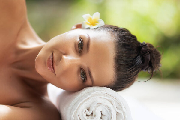 Smiling Asian woman resting at outdoor spa, wrapped in towel with frangipani flower in hair. Perfect beauty and wellness image for spa services, skincare ads, and self-care promotion.