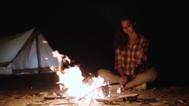 Genç bir kadın, arka planda yumuşak bir çadırla, kumlu bir kamp alanında, çatırdayan bir kamp ateşinin yanında oturuyor. Açık havada yaşamak, rahatlamak ve kamp maceraları için mükemmel..