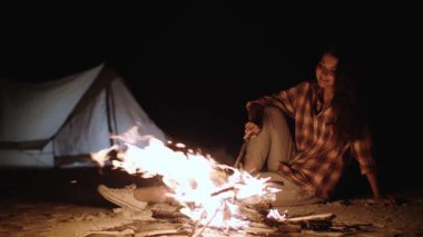 Huzurlu genç bir kadın, arka planda parlayan bir çadırla kumlu bir kamp alanındaki parlak bir kamp ateşinin yanında dinleniyor. Açık havada yaşamanın ve huzur içinde kaçmanın güzelliğini yakalar..