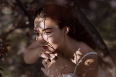 Ağaç dallarının altında dinlenen, yüzü benekli güneş ışığıyla aydınlatılmış, yumuşak yüzlü genç bir kadının sanatsal portresi. Nazik ve karamsar bir bahar anı güzellik kavramları için mükemmel..