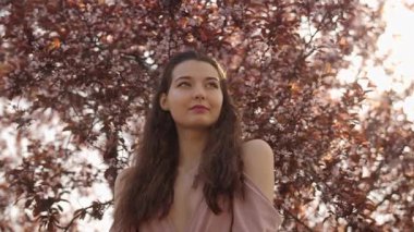 Yumuşak pembe elbiseli uzun saçlı genç kadın sakura çiçeklerinin altında poz veriyor. Güzellik, moda ve ilkbahar yaşam tarzı kavramları için yüksek kaliteli yavaş çekim görüntüleri.