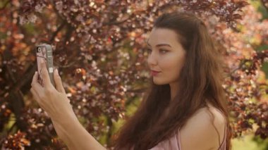 Beyaz kadın, kiraz çiçeklerinin altında sıcak, doğal bir ışıkla selfie çeker. Yaşam tarzı, güzellik, sosyal medya ve akıllı telefon teknolojisi tanıtımı için mükemmel..