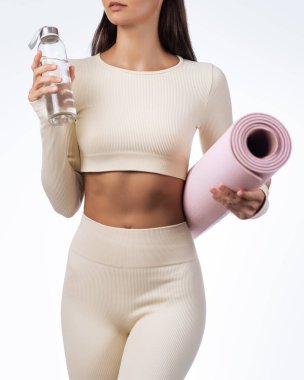 Kremalı elbisesi olan, elinde pembe yoga minderi ve cam su şişesi tutan tonlarca sporcu kadın. Stüdyo çekimi spor, güzellik veya sağlıklı yaşam tarzı reklamcılığı ve promosyon tasarımı için idealdir..