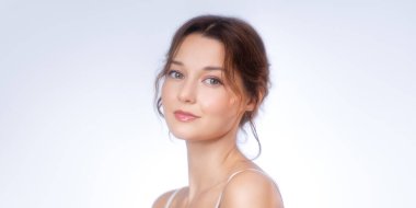 Doğal, ışıl ışıl makyajlı zarif bir genç kadının stüdyo portresi. Nazikçe gülümser ve yavaşça kameraya bakar. Sağlıklı, parlak cildini gösterir. Güzellik, cilt bakımı, kozmetik için ideal.