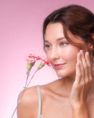Genç beyaz bir kadının zarif dikey portresi nazikçe yüzüne dokunuyor ve pembe karanfiller kokluyor. Cilt bakımı, çiçek güzelliği ya da romantik kozmetik ürünleri promosyonları için ideal..