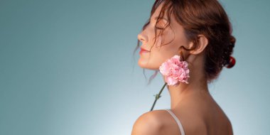 Minimalist ve şiirsel bir stüdyo portresi. Gözleri kapalı, kulağının arkasında pembe bir karanfil tutan genç bir kadın. Yumuşak mavi arka plan ve nazik yan ışık onun doğal güzelliğini vurgular.