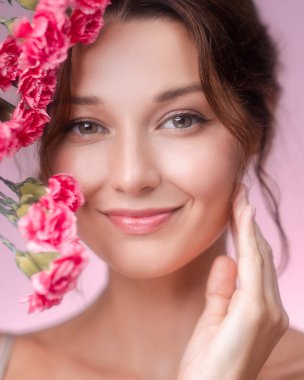 Canlı pembe çiçeklerle çevrili genç bir kadının gülümseyen parlak ve neşeli yakın plan portresi. Işıl ışıl cildi ve doğal makyajı pastel pembe arka planda parlıyor.