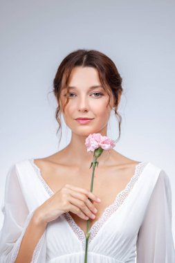 Narin beyaz elbiseli, elinde pembe bir karanfil çiçeği tutan güzel bir kadın. Zarif güzellik ve cilt portresi, kaplıca, moda, yaşam tarzı, reklamcılık ve yüksek kaliteli tanıtım için mükemmel.