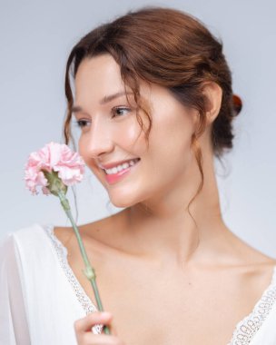 Beyaz bir kadının soluk pembe bir çiçeğe gülümseyen doğal güzelliği. Minimalist stüdyo geçmişi, temiz cilt bakımı, kozmetik reklamlar ve kadın yaşam tarzı görselleri için mükemmel..