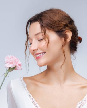 Pembe bir karanfille gülümseyen, beyaz bluz giyen, güzellik, cilt bakımı, çiçekli yaşam tarzı ve sağlık temalı promosyon tasarımları için mükemmel genç beyaz bir kadının parlak stüdyo portresi..