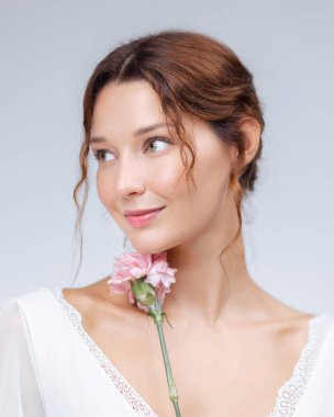 Beyaz dantel elbiseli, elinde pembe çiçekle şık bir genç kadının yakın plan fotoğrafı. Işıl ışıl teni ve nazik görünüşü cilt bakımı, kozmetik ürünleri ve zarif güzellik kampanyaları için idealdir..