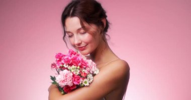 Kafkasyalı genç bir kadının pastel arka planda yumuşak bir gülümsemeyle pembe bir buketi kucakladığı yavaş çekim videosu. Güzellik, parfüm, cilt bakımı ve bahar reklamları için mükemmel..