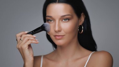 Kusursuz derisi olan güzel genç bir kadın kameraya güvenle bakarken yumuşak bir fırça kullanarak makyaj yapıyor. Güzellik, kozmetik ve cilt bakımı için ideal.. 