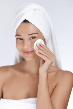 Kafasında beyaz bir havlu olan beyaz bir kadın, yanağını pamukla nazikçe temizliyor. Cilt bakımı, kaplıca, güzellik ve kozmetik kavramları için harika bir görüntü..
