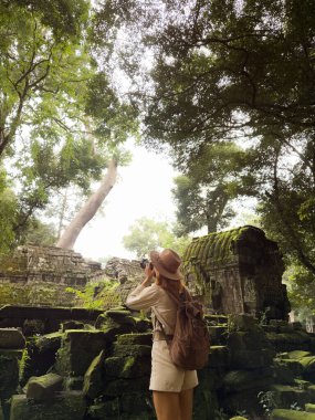 Kamboçya 'daki Ta Prohm tapınağının kalıntılarının fotoğraflarını çeken sırt çantalı ve şapkalı genç bir gezgin. Orman ve yosun kaplı taşlarla çevrili. Seyahat, kültür, macera ve turizm tanıtımı için mükemmel..