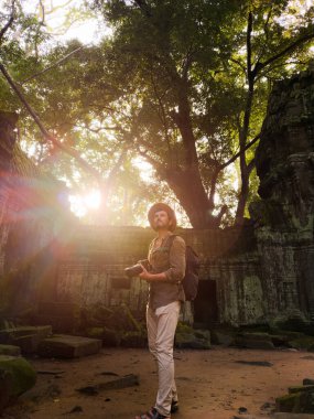 Güneş doğarken Angkor, Kamboçya 'daki Ta Prohm tapınağının kalıntılarını fotoğraflayan sırt çantalı ve kameralı bir erkek gezgin. Seyahat fotoğrafçılığı, macera, yaşam tarzı ve kültürel miras tanıtımı için mükemmel..