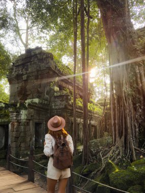 Kamboçya 'daki Ta Prohm tapınağı kalıntılarını araştıran sırt çantalı kadın turistin arkası. Antik taş duvarların üzerindeki devasa ağaç kökleri ile ünlüdür. Seyahat, kültür, macera ve miras tanıtımı için ideal.