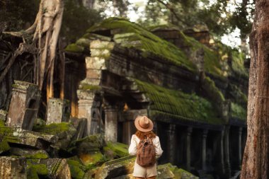 Kamboçya 'nın Angkor kompleksindeki Ta Prohm kalıntılarını ziyaret eden şapka ve sırt çantalı kadın turist, orman ve yosunlarla çevrili. Seyahat, yaşam tarzı, kültür ve macera tanıtım kullanımı için mükemmel.