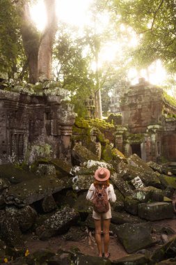 Kamboçya 'nın Angkor kompleksindeki ormanla kaplı Ta Prohm tapınağının kalıntıları arasında yürüyen sırt çantası ve şapka takan genç bir kadın turist. Seyahat, yaşam tarzı, kültür ve macera tanıtım kullanımı için harika.