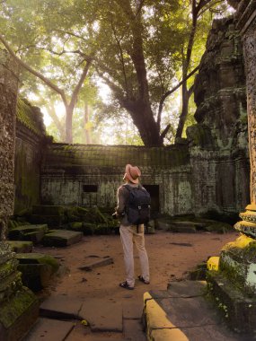 Kamboçya 'nın Angkor kompleksindeki Ta Prohm kalıntılarını araştıran ve fotoğraflayan sırt çantalı adam, orman ve gün ışığıyla çevrili. Seyahat, yaşam tarzı ve kültür tanıtımı için harika..
