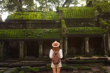 Kamboçya 'nın Angkor kentindeki yosun kaplı Ta Prohm tapınağını araştıran şapka giyen sırt çantalı kadın. Seyahat, yaşam tarzı, turizm, tarih, macera ve kültür mirası kullanımı için harika.