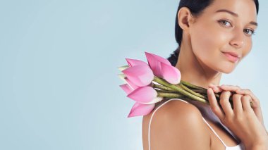 Omzunda pembe nilüfer çiçekleri tutarken gülümseyen zarif genç bayanın yakın çekimi. Parfüm, spa, cilt bakımı ya da kozmetik promosyonları için mükemmel bir stüdyo portresi..