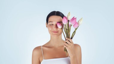 Gülümseyen kadın yüzünde pembe nilüfer tomurcukları tutarak doğal güzellik ve cilt bakımını sembolize ediyor. Spa tedavileri, sağlık promosyonları, güzellik ürünü reklamları ve lüks için ideal.