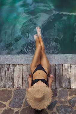 Siyah bikinili genç bir kadın tahta bir şapkayla güvertede uzanıyor, ayaklarını yüzme havuzuna daldırıyor. Yaz tatili, dinlenme, seyahat ve boş zaman için yüksek kaliteli yaşam tarzı konsepti.