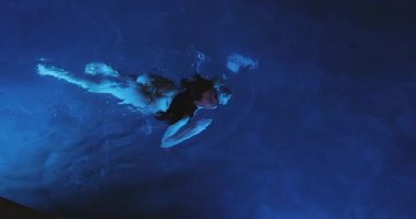 Mavi ışıklı bir havuzda su altında yüzen bir kadın zarif bir şekilde suda süzülüyor ve etrafında dalgalanmalar yaratıyor. Bir kadın zarif bir şekilde suyun altında yüzer, mavi ışıkla aydınlatılır, dingin bir hava yaratır.