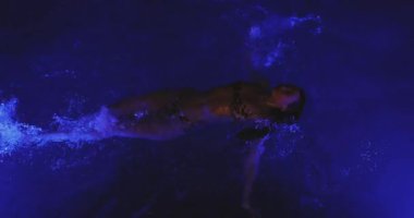 Yukarıdan yakalanan bir kadın, gece vakti parlak mavi bir havuzda zahmetsizce süzülür. Süper yavaş çekim etkisi rüya gibi atmosferi güçlendirir. Suyun hipnotik parıltısını vurgular.. 