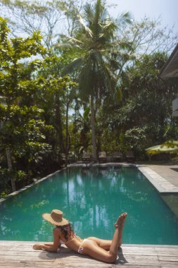 Beyaz bikinili ve hasır şapkalı bir kadın, yemyeşil bir otelin havuzunda dinleniyor. Huzurlu ortam lüks, rahatlama ve egzotik tatil titreşimleri temaları vurguluyor.