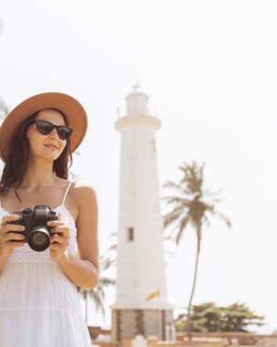 Şapkalı ve güneş gözlüklü bir kadın Sri Lanka 'daki ikonik Galle Fort deniz fenerinin önünde kamera tutuyor. Palmiye ağaçlarının olduğu güneşli tropik sahne macera ve seyahat fotoğrafçılığını yansıtıyor..