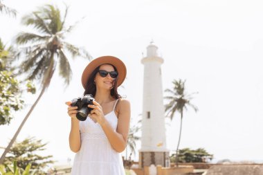 Sri Lanka 'da palmiye ağaçları olan Galle Deniz Feneri' nin yanında kamerayla gülümseyen hasır şapkalı ve güneş gözlüklü genç kadın, tropik tatil, gezi, yaşam tarzı fotoğrafçılığı ve macera kavramı.