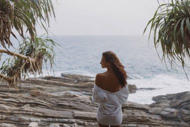 Beyaz gömlekli genç bir kadın kayalık Sri Lanka sahilinden okyanus dalgalarını seyrediyor. Kavramsal yaşam tarzı, seyahat, rahatlama ve özgürlük teması, refah, promosyon veya yayın için ideal.