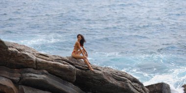 Kırmızı bikinili bir kadın okyanus dalgalarını gören kayalık bir uçurumda güven içinde oturuyor. Macera, seyahat ve sahil yaşam tarzı promosyonları için mükemmel, özgürlüğü ve rahatlamayı sembolize ediyor..