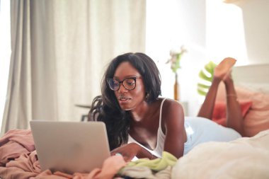 Bilgisayarla çalışırken yatakta uzanan siyah kadının portresi.