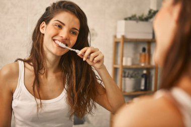 Genç bir kadın aynada dişlerini fırçalar.