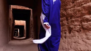 Mhamid, Fas 'taki Kasbah' ın içinde mavi gandoura ve türban giyen geleneksel giyimli bir Bedevi adam yürüyor. Güney Fas 'ın otantik kırsal manzarası. 4k.  