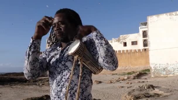 アフリカのパーカッションミュージック セネガル人男性は スティックで伝統的な小さな砂丘のドラムを演奏しています モロッコのエサウイラのビーチで屋外ストリートミュージック 4Kについて — ストック動画