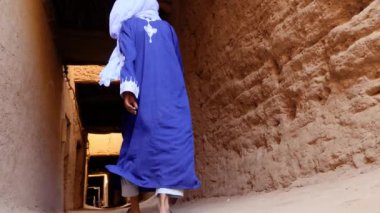 Geleneksel giyinmiş Faslı bir Bedevi adam mavi gandoura giyiyor ve Mhmaid, Fas 'ta Kasbah' ın içinde türban yürüyor. Güney Fas 'ın otantik kırsal manzarası. 4k. 