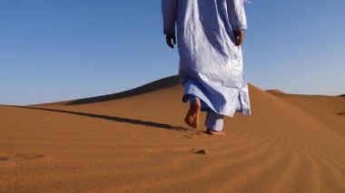 Geleneksel olarak giyinmiş Faslı bir adam mavi gandoura, djellaba ve türban giyerek kum tepeciklerinde yürür, Erg Chegaga Çölü, Fas. Güney Fas 'ın otantik çöl sahnesi. 4k. 