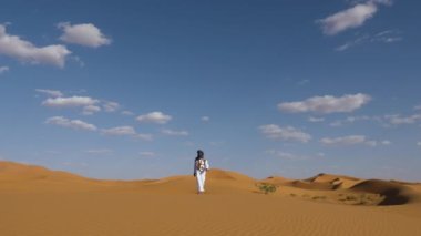 Geleneksel olarak beyaz gandoura, djellaba ve türban giyen Faslı bir adam kum tepeciğinde yürüyor, Erg Chebbi Çölü, Fas. Güney Fas 'ın otantik çöl sahnesi. 4k.  