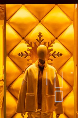 Manken kışlık giysiler giyer, vitrinde sergilenir. Sarı arka plan ve ahşap kar tanesi süslemesi vardır.