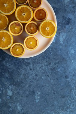 Dilimlenmiş portakallar ve kan portakalları pembe bir tabağa dizilir ve dokulu mavi arkaplana karşı canlı bir kontrast oluşturur.