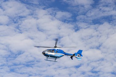 Bulutlu bir havada uçan mavi ve beyaz helikopter, hava ulaşımı ve hizmetleri sağlar.