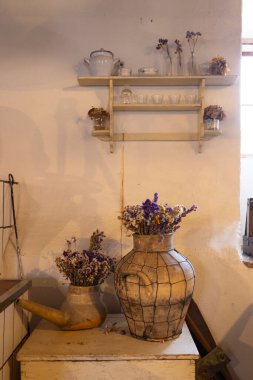 Eski vazolarda kurumuş çiçeklerle süslenmiş taşralı bir mutfak ve eski çanak çömlekle dolu bir raf, rahat ve nostaljik bir atmosfer yaratıyor.